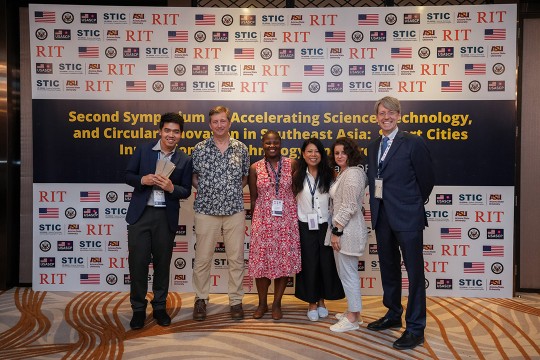 a group of six students and adults stand in front of a sign bearing logos from R I T and other companies.