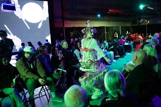 people sit on both sides of a fashion show runway where a person in a dress in clown styling walks down the center.