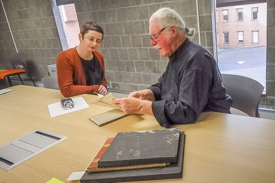 A man with white hair and glasses shows an old document to a woman wearing glasses and a red cardigan, as they sit at a table with archival materials in a modern office setting.