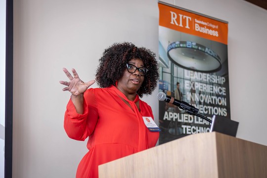 A woman in a red blouse speaks at a podium with a microphone, gesturing with her hand. A banner behind her reads RIT Saunders College of Business with keywords like leadership and innovation.