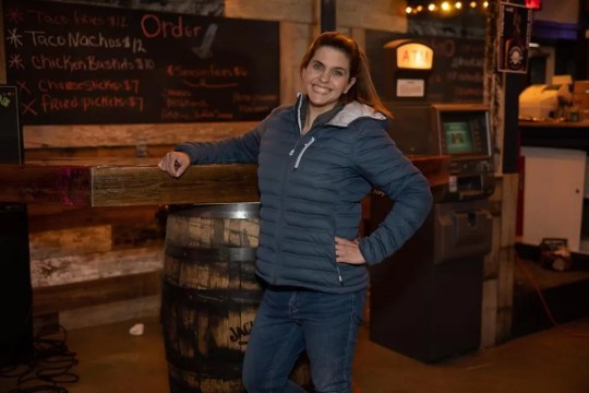 A woman leans against a barrel in a lively bar, looking relaxed and happy.