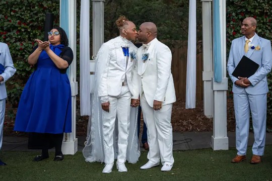 Newlywed couple in matching white suits share a kiss at their wedding ceremony, holding hands under an outdoor arch, while an officiant and a sign language interpreter stand nearby.