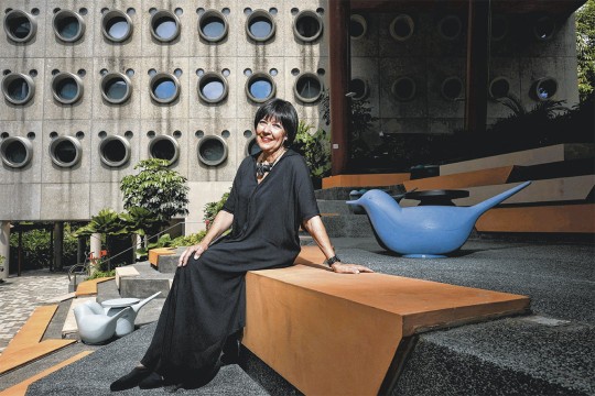 A woman in a black dress sits on a geometric bench in an outdoor space with modern architecture featuring circular windows. She smiles at the camera, surrounded by sculptural blue bird-shaped seats and greenery.