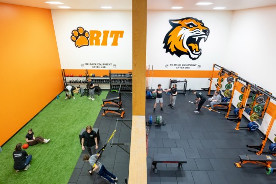 Split view of a fitness center at Rochester Institute of Technology (RIT), with one side featuring turf and functional training equipment, and the other side equipped with squat racks and barbells. Students exercise in both areas, with the RIT logo and tiger mascot on the walls.