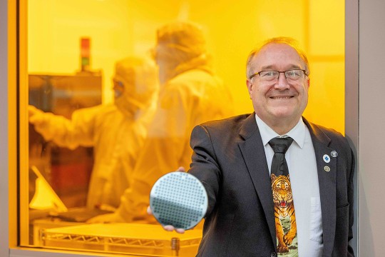 Robert Pearson stands in front of the MicroE clean room. Robert is in a suit, sporting a tie with a realistic tiger on it and several RIT pins, and holds a semiconductor out toward the camera.