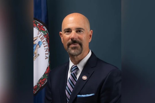 Man in suit and tie with Virginia flag partially behind him. 