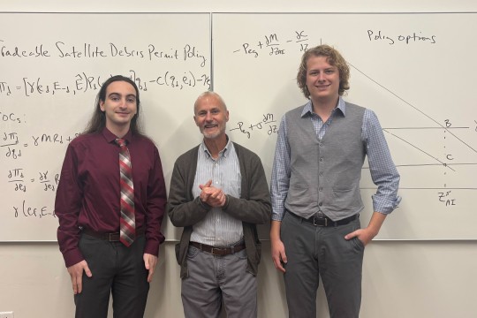 three men stand in front of a whiteboard in a classroom smiling at the camera.