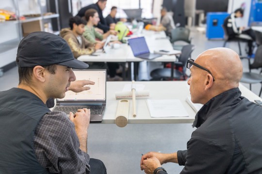 a student in a black hat speaks with a man sitting to his right, gesturing near a laptop screen.