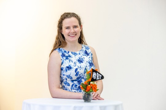 a college age female in a blue and white floral dress stands in front of a white table holding a small bouquet of orange flowers with an R I T flag nestled within them.