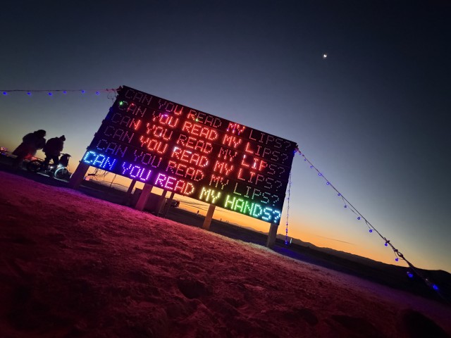 Art installation 'Can You Read My Lips?' for Burning Man 2026, shown here at dawn in Nevada’s Black Rock Desert. Five rows of the 15-by-7-foot LED sign repeat a question Deaf people are asked their whole lives, with an ask on the last line.
