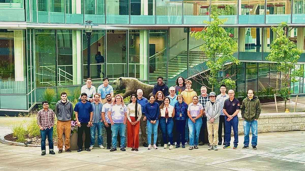 a group of about 20 people poses for a photo outdoors in front of a tiger statue and a glass building.