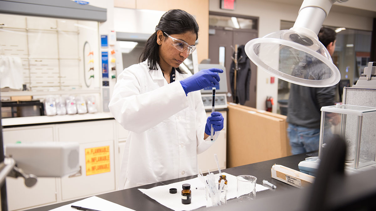 a researcher wearing safety glasses and a white lab coat uses a syringe to measure a dark liquid.