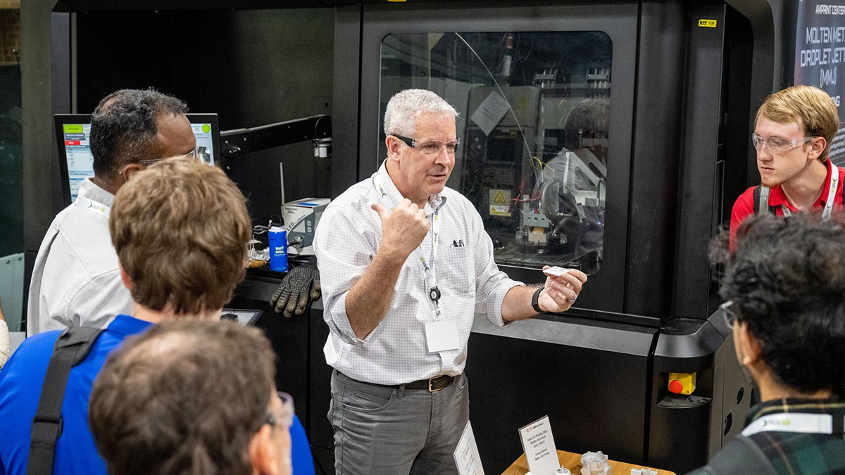 a man wearing safety glasses in a lab discusses the equipment with visitors.
