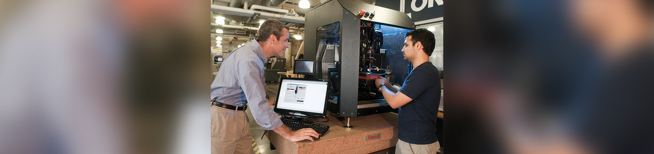 Denis standing over a laptop while peering into som 3d printer with another person working beside him