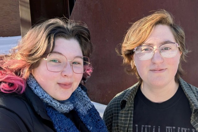 two people smile for the camera in front of an iron wall.