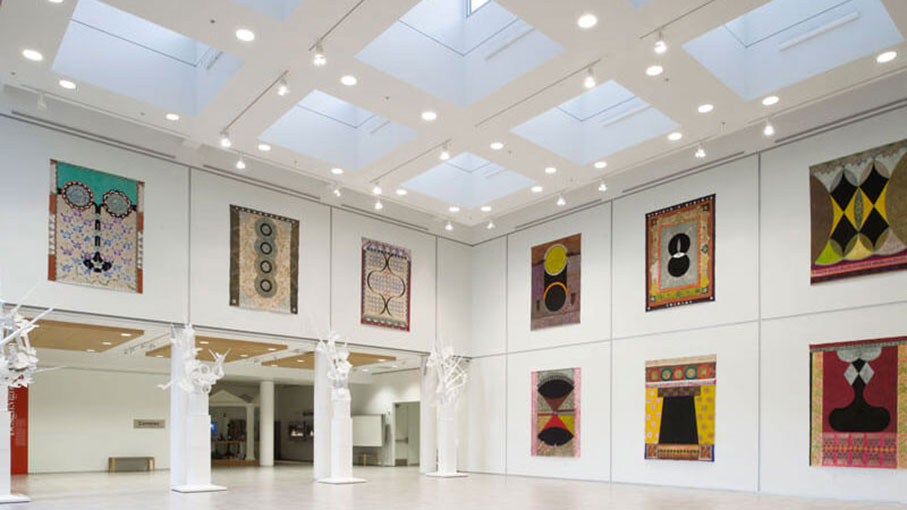 A brightly lit modern art gallery displaying colorful quilts and sculptures under skylights.