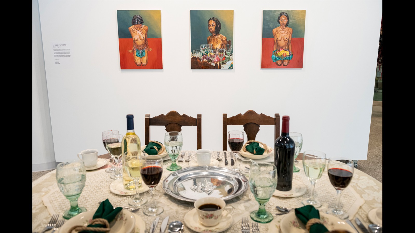Paintings are mounted on a wall behind a table set with a cloth, wine and glasses.