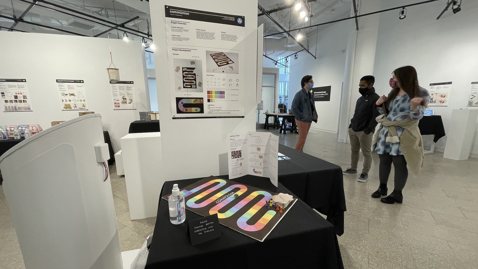 A board game design sits in the foreground of a photo while students in the background talk.