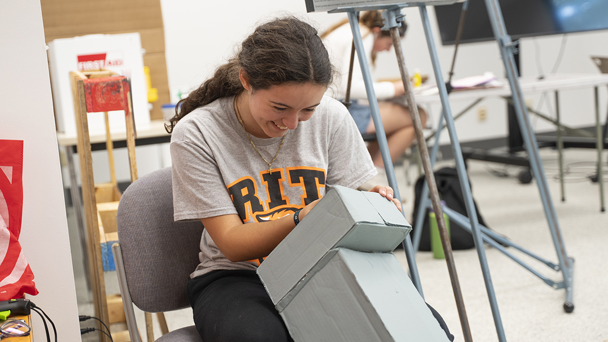 A student smiles as they work on a robot creation made of cardboard.