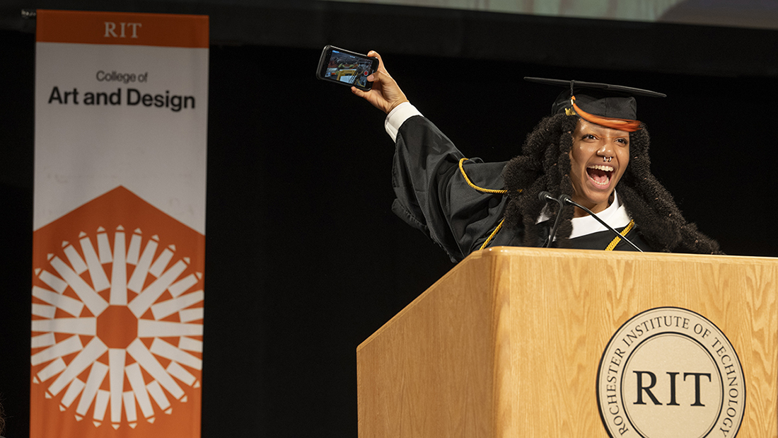 A student in regalia take a selfie at the lectern during commencement.
