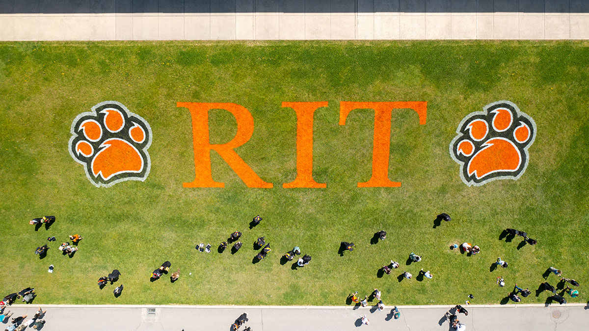 An aerial view of a campus lawn with 'R I T' painted in orange letters and two large orange tiger paw prints.
