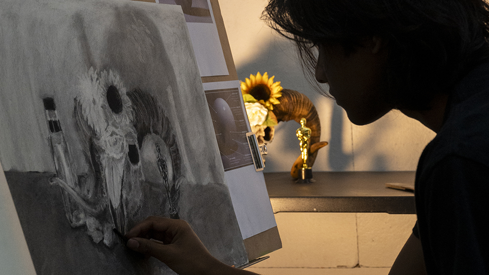 A silhouette of a student in front of an easel, drawing a still life.