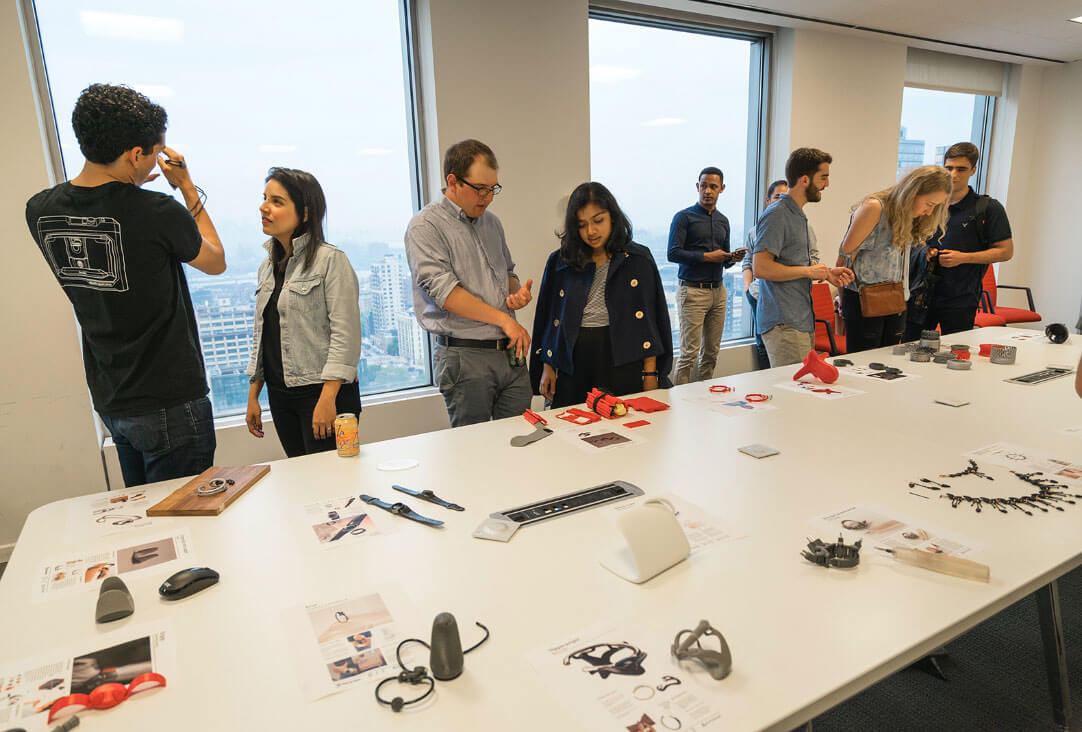 Nine people in a room looking at items that have been 3D printed.