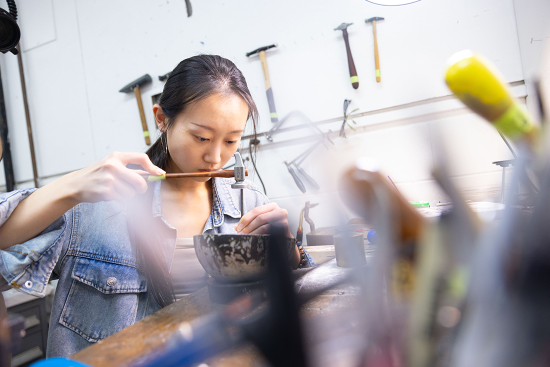 An RIT metals and jewelry design student hammers an object on a table.