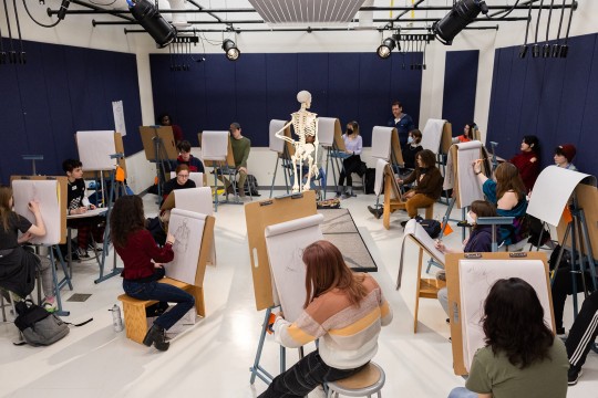 students in a drawing class sit at easels and draw a skeleton posing in the center of the studio.