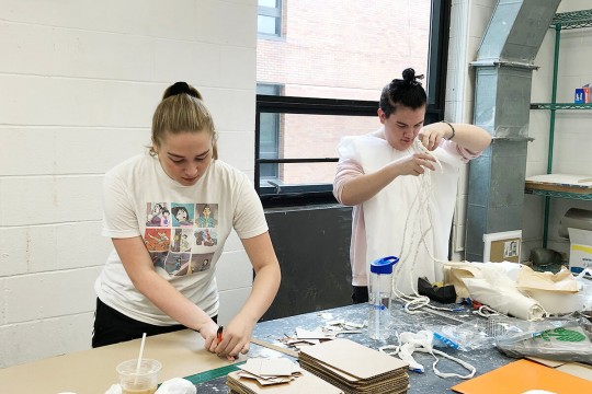 two students working on sculpture projects at a large work table.