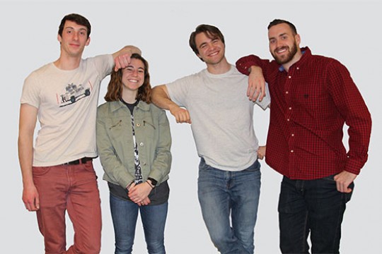Four members of the Gaia team stand together, posing for a photo by leaning against each other's shoulders with their elbows. James has his elbow on Kerri's head to accentuate their height difference.