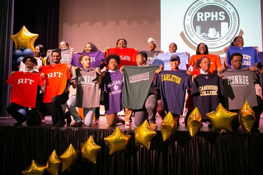 A group of graduating students from Rochester Prep High School stand on a stage and hold up t-shirts from the colleges they have been accepted into for the fall.