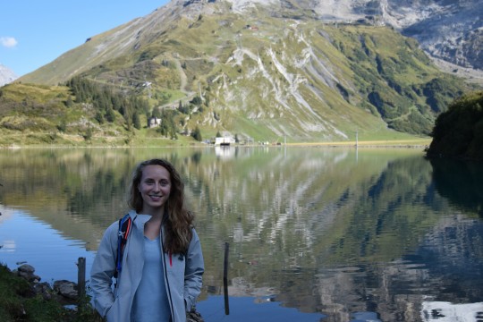 Scholl posing in front of lake with large mountains behind her