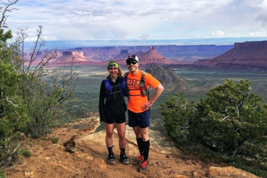 Couple in hiking gear stands on ledge with canyon in background.