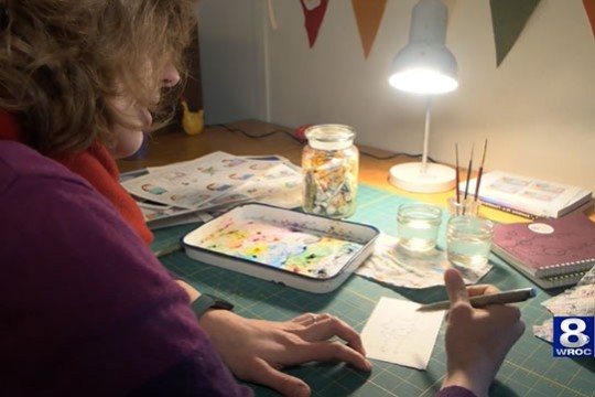 artist drawing a cartoon while seated at a desk with jars of water and a paint tray.