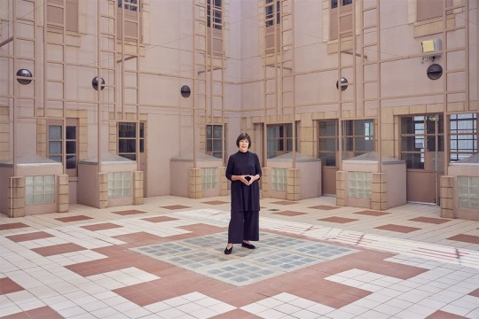 woman dressed in all black standing in a large room with a tiled floors and scaffolding accents on the walls.
