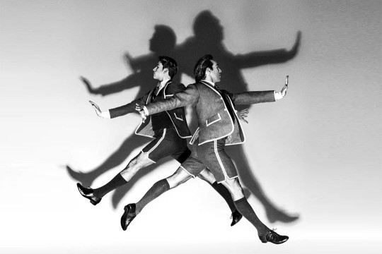 black and white image of two male ballet dancers wearing a suit jacket and shorts with white trim leaping away from each other with their arms spread out.