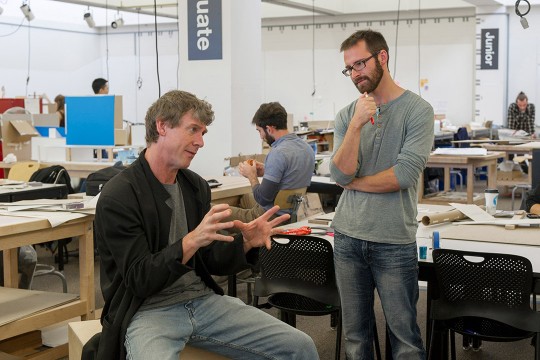 seated professor talking to standing student.