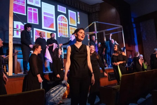 a woman in a black dress performs on stage with other show members behind her.