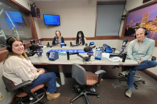 Four people wearing headphones sit at a table in a radio talk studio: a woman front left has long brown hair and is wearing a beige shirt, blue jeans and brown boots; a man front right has short dark hair and is wearing a light green button-down shirt, blue jeans and sneakers; a woman back left has long blonde hair and is wearing a black zip-up fleece over a grey t-shirt; a woman back right has long black and green dreadlocks and is wearing glasses and a pink shirt.