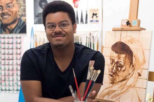 a man sits next to a painting of a female in an art studio.