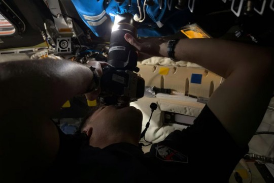 An astronaut inside a spacecraft holding a camera up to a window with a cloth covering. The lens is pushed through a hole in the covering