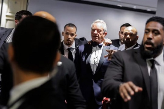 R F K junior is flanked by security officers in a white room.