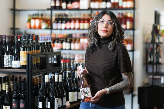 Tanvi Asher, a woman with dark curly hair and steel rimmed glasses, holds a bottle of Rose with a colorful label. In the background, shelves and tables full of wine sit in Bonesteel Wines and Spirits