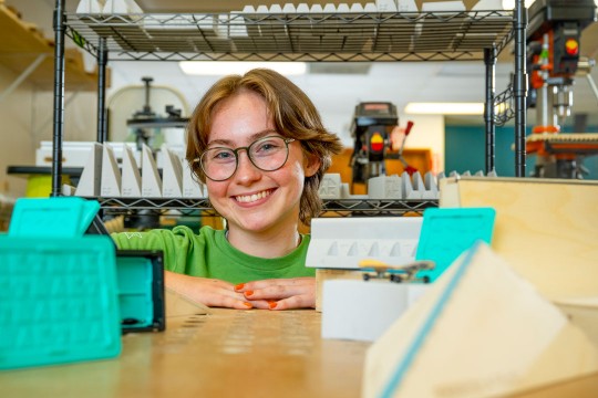 a collage age person with short hair and glasses looks at the camera through a shelving unit