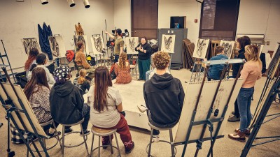 a group of students sits in a circle on stools next to their drawings.