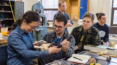 student holds a wooden art piece in a clamp while another person takes a photo of it.