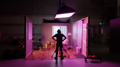 a student stands with their hands on their hips looking at a scene of a small living room in pink lighting.