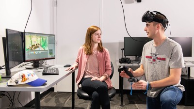 two students in a V R lab, with one wearing V R goggles and holding handheld devices.