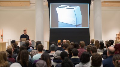 a speaker addresses a full room, with a toaster design on a projector screen.